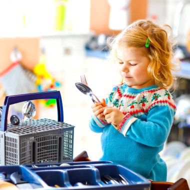 Küçük tatlı kız mutfakta bulaşık makinesiyle yardım ediyor. Bıçakları, çatalları, kaşıkları, çatalları ayıran mutlu sarışın çocuk. Bebek anne ve babaya yardım ederken eğleniyor..