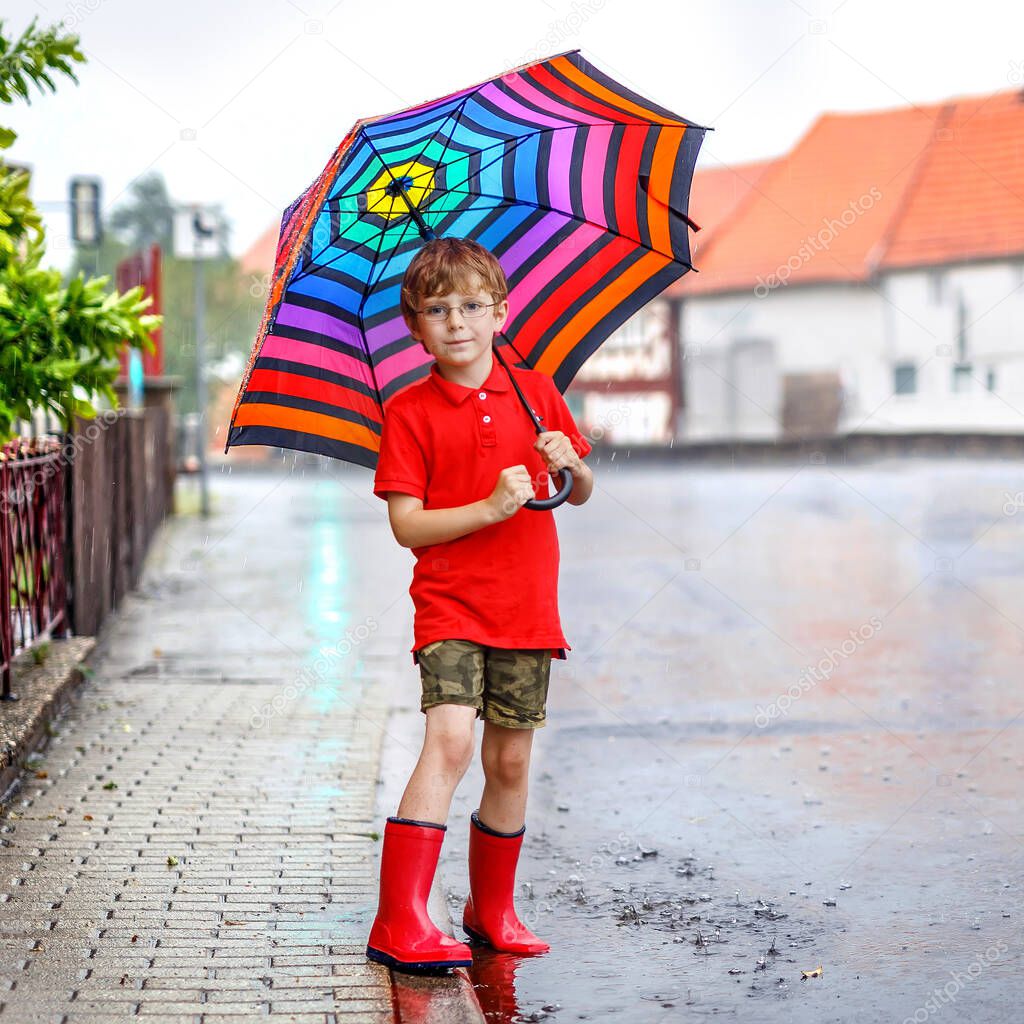 Kid boy wearing red rain boots and walking with colorful umbrella on city street. Child with glasses on summer day. happy kid during heavy summer shower rain.