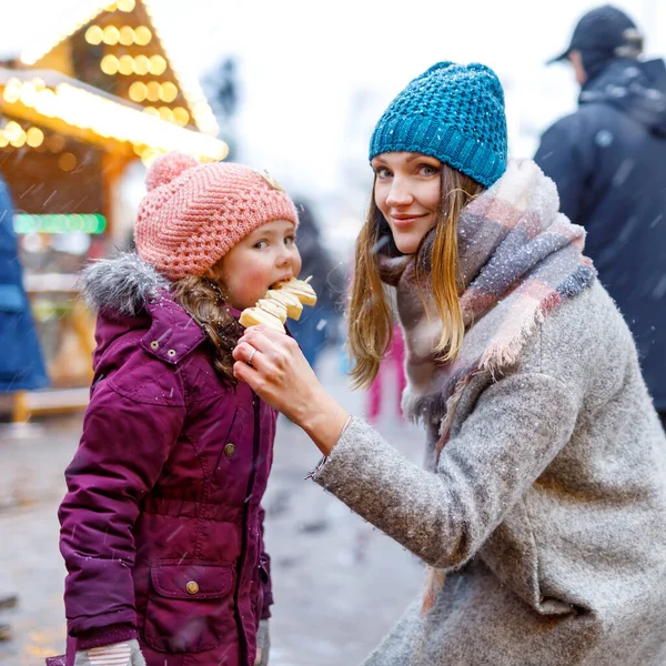 Young mother and daughter eating white chocolate covered fruits on skewer on traditional German Christmas market. Happy girl and woman on traditional family market in Germany, Munich during snowy day. — Stock Photo, Image