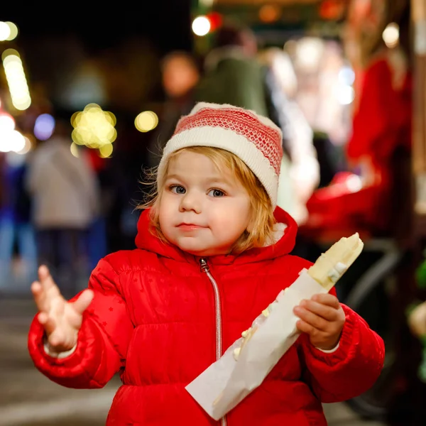 小さな女の赤ちゃん、かわいい子がバナナを食べては、チョコレート、marshmellows、ジンジャーブレッド、ナッツと甘いスタンド近くカラフルなスプリンクルで覆われています。ドイツのクリスマス マーケットで幸せな幼児. — ストック写真