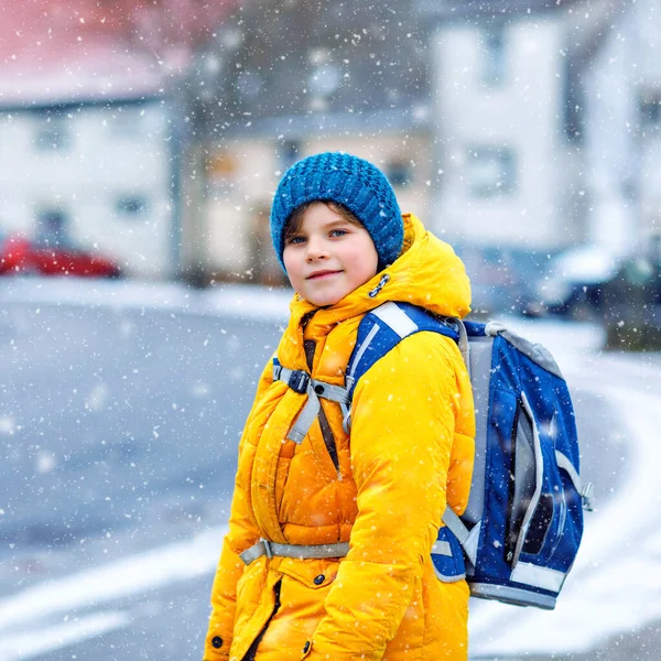 Menino da escola de classe primária a caminhar para a escola durante a queda de neve. Criança feliz se divertindo e brincando com a primeira neve. Estudante com jaqueta amarela e mochila em roupas coloridas de inverno . — Fotografia de Stock