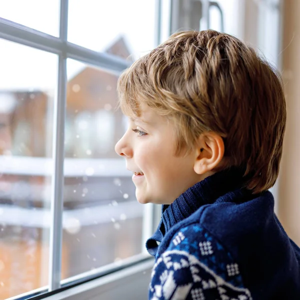 Niño adorable feliz sentado cerca de la ventana y mirando hacia fuera en la nieve en el día de Navidad o la mañana. Niño sonriente fascinado con nevadas y grandes copos de nieve — Foto de Stock