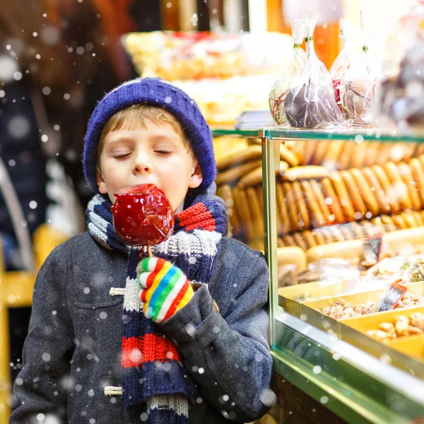 Pequeno menino bonito perto doce stand com pão de gengibre e nozes. Criança feliz comendo em maçã coberta de açúcar vermelho. doce tradicional no mercado de Natal alemão.. — Fotografia de Stock