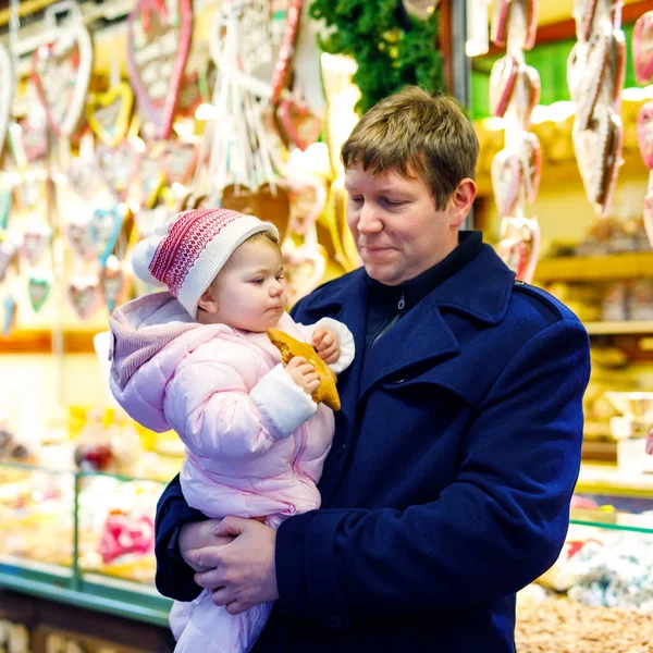 Middle aged father holding baby daughter near sweet stand with gingerbread and nuts. Happy family on Christmas market in Germany. Cute girl eating cookie called "Lebkuchen". Celebration xmas holiday. — Stock Photo, Image