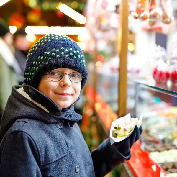 Маленький мальчик, милый ребенок ест бананы, покрытые шоколадом, мармеллоу и красочные брызги возле сладкого стенда с пряничным хлебом и орехами. Счастливый мальчик на рождественской ярмарке в Германии . — стоковое фото