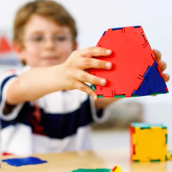 Маленький мальчик в очках играет с пластмассовым набором элементов в школе или детском саду. Счастливый ребенок строит и создает геометрические фигуры, учится игре и геометрии. — стоковое фото
