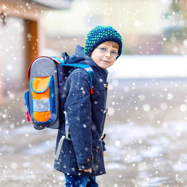 Kleiner Schulbub der Grundschule auf dem Weg zur Schule. Glückliches Kind, das Spaß hat und mit dem ersten Schnee spielt. Student mit Rucksack in bunter Winterkleidung. Schulgebäude. — Stockfoto