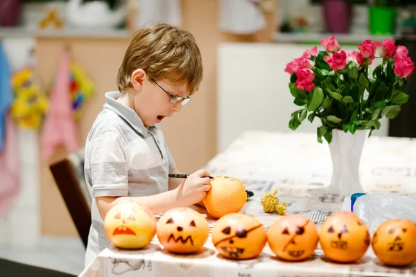 Little kid boy making Jack-o-lantern with drawing scary faces on mandarine, tangerine or clementine. Happy child making preparation for Halloween party at home, indoors — Stock Photo, Image