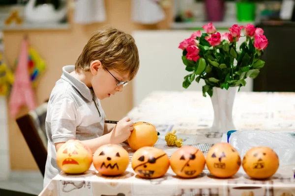 Little kid boy making Jack-o-lantern with drawing scary faces on mandarine, tangerine or clementine. Happy child making preparation for Halloween party at home, indoors — Stock Photo, Image