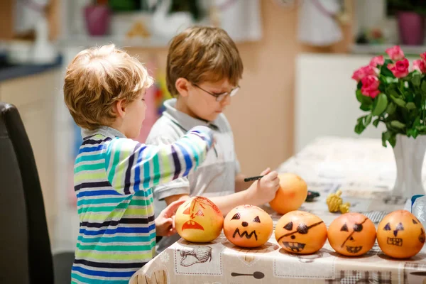 Két kisfiú Jack-o-lámpást készít ijesztő arcokkal mandarinra, mandarinra, mandarinra vagy klementinra. Boldog gyerekek testvérek és ikrek készülnek Halloween party otthon, beltérben — Stock Fotó