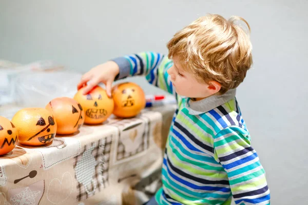 Little kid boy making Jack-o-lantern with drawing scary faces on mandarine, tangerine or clementine. Happy child making preparation for Halloween party at home, indoors — Stock Photo, Image
