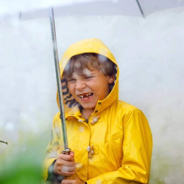 Beautiful little kid boy on way to school walking during sleet, heavy rain and snow with an umbrella on cold day. Happy and joyful child in colorful yellow coat fashion casual clothes — Stock Photo, Image