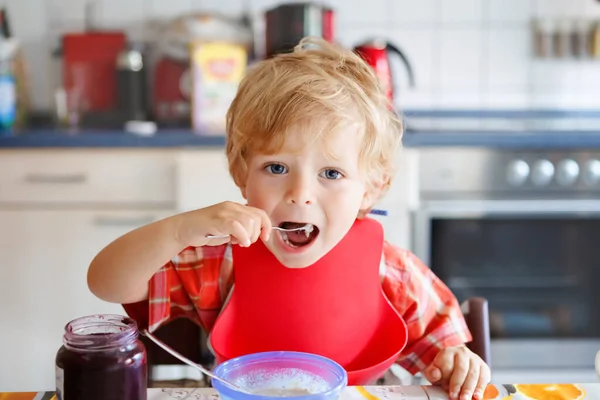 Entzückender kleiner Junge, der gesunde Schweinefleisch vom Löffel zum Frühstück mit Beerenmarmelade isst. niedliches fröhliches Baby im bunten Pyjama sitzt in der Küche und lernt mit dem Löffel. — Stockfoto