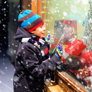 Noel pazarındaki şirin küçük okul çocuğu. Moda kışı kıyafetleri içinde, hediyelerle süslenmiş vitrin alışverişi yapan neşeli çocuk, xmas ağacı. Kar yağıyor, kar yağıyor..