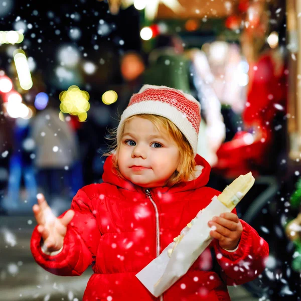 Bambina, graziosa bambina che mangia banane ricoperte di cioccolato, marshmellows e colorate spruzzate vicino al dolce stand con pan di zenzero e noci. Buon bambino al mercatino di Natale in Germania . — Foto Stock