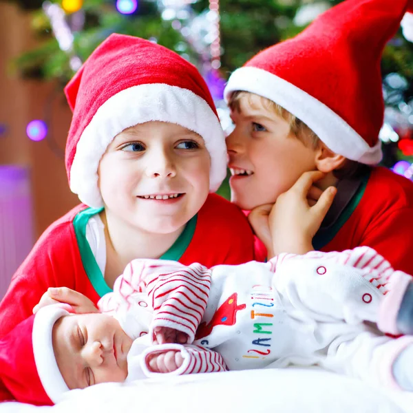 One week old newborn baby girl and two siblings kid boys in Santa Claus hats near Christmas tree with colorful garland lights on background. Close-up of three children, happy family celebrating Xmas. — Stock Photo, Image