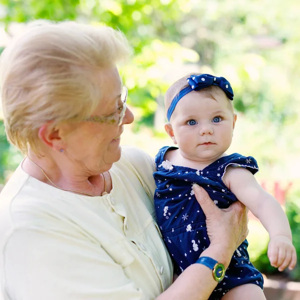 Χαριτωμένο κοριτσάκι με γιαγιά την καλοκαιρινή μέρα στον κήπο. Ευτυχισμένη ηλικιωμένη γυναίκα που κρατά το χαμογελαστό παιδί στο χέρι.. — Φωτογραφία Αρχείου