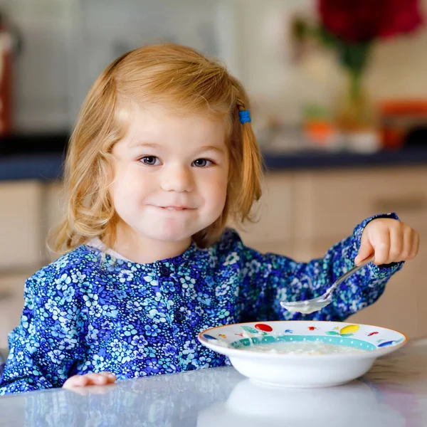 Schattig peutermeisje dat gezonde ontbijtgranen met melk eet. Schattig gelukkig baby kind in kleurrijke kleren zitten in de keuken en plezier hebben met het bereiden van haver, granen. Binnenshuis thuis — Stockfoto