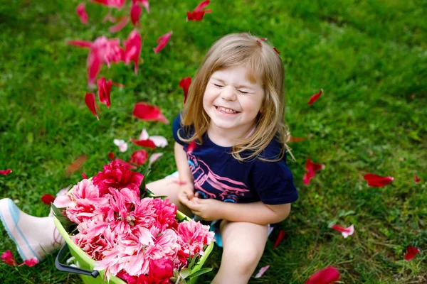 Carino adorabile bambina con un sacco di fiori di peonia rosa nel secchio. Felice bambino sorridente in giardino domestico nella calda giornata primaverile o estiva. — Foto Stock