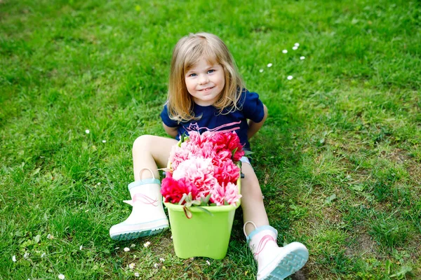 バケツにピンクの牡丹の花がたくさんあるかわいいかわいい幼児の女の子。暖かい春や夏の日に国内庭で幸せな笑顔の子供. — ストック写真