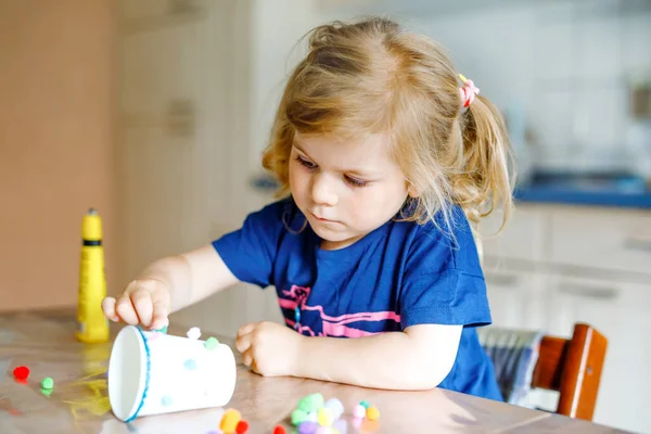 Niña pequeña haciendo linterna artesanal con vasos de papel, pompones de colores y pegamento durante la enfermedad pandémica de cuarentena coronavirus. Feliz niño creativo, educación en casa y guardería en casa con los padres — Foto de Stock