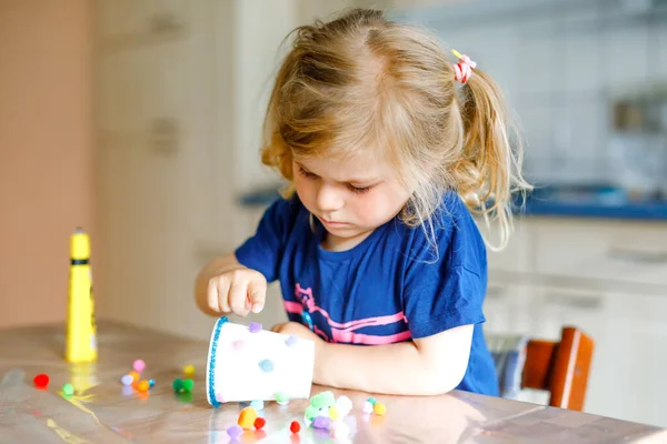 Egy kislány, aki papírpoharakkal, színes pomponokkal és ragasztóval készíti a kézműves lámpást a világméretű koronavírus karantén alatt. Boldog kreatív gyermek, otthoni tanulás és otthoni napközi a szülőkkel — Stock Fotó