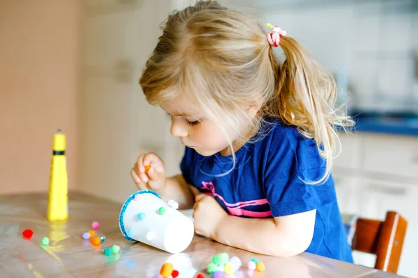 Pequena menina fazendo lanterna artesanal com copos de papel, pompons coloridos e cola durante a pandemia coronavírus quarentena doença. Criança criativa feliz, educação em casa e creche em casa com os pais — Fotografia de Stock