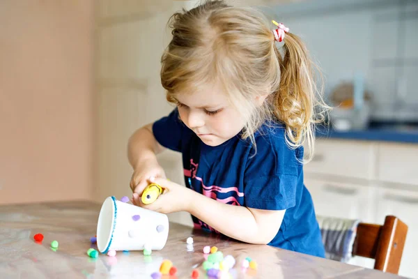 Kleine peuter meisje maken ambachtelijke lantaarn met papieren bekers, kleurrijke pompons en lijm tijdens pandemische coronavirus quarantaine ziekte. Gelukkig creatief kind, thuisonderwijs en thuisopvang met ouders — Stockfoto