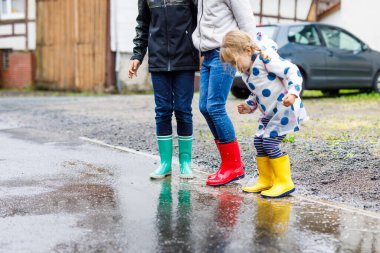 Kırmızı, sarı ve yeşil çizme giyen ve yağmurluk sırasında yürüyen üç çocuk, küçük bir kız ve iki çocuk. Mutlu kardeşler gölete atlıyor. Dışarıda eğleniyor, aktif bir aile.