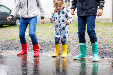 Kırmızı, sarı ve yeşil çizme giyen ve yağmurluk sırasında yürüyen üç çocuk, küçük bir kız ve iki çocuk. Mutlu kardeşler gölete atlıyor. Dışarıda eğleniyor, aktif bir aile.