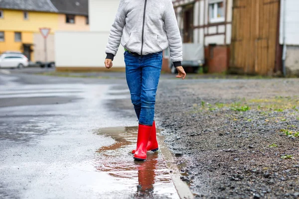 Detailní záběr chlapce v červených holínkách a chůze během deště a deště v deštivém oblačném dni. Dítě v barevných neformálních šatech skákající do kaluže. Bavíte se venku, zdravé aktivity dětí — Stock fotografie