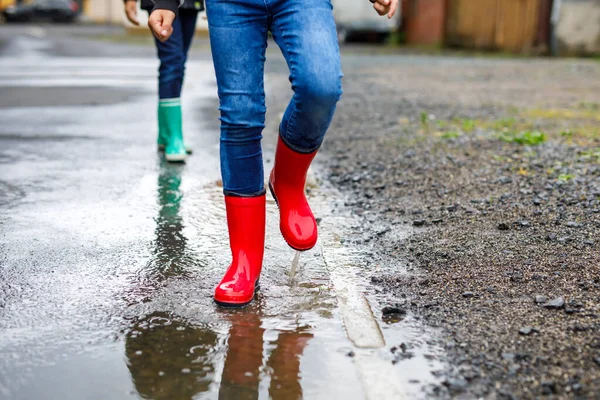 Primer plano de las piernas de dos niños niños con botas de lluvia rojas y verdes y caminando durante el aguanieve y la lluvia en el día nublado lluvioso. Niños saltando al charco. Divertirse al aire libre, actividad saludable para niños — Foto de Stock