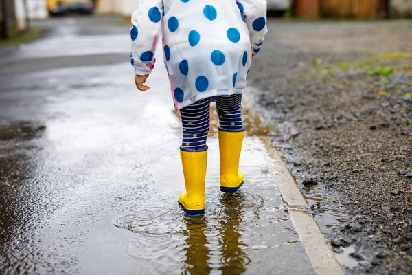 Close-up of little toddler girl wearing yellow rain boots and walking during sleet on rainy cloudy day. Cute child in colorful clothes jumping into puddle, splashing with water, outdoor activity — Stock Photo, Image