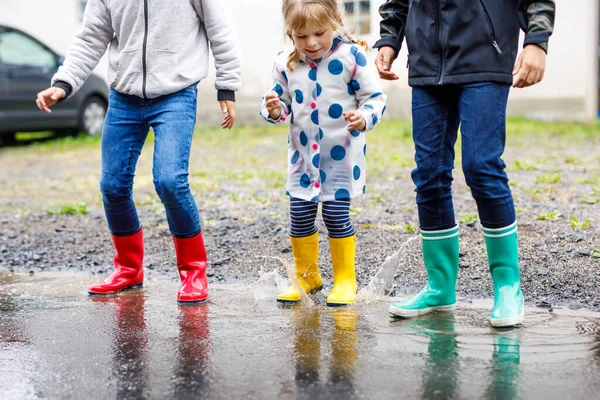 Närbild av tre barn, småbarn flicka och två barn pojkar bär röda, gula och gröna regnstövlar och promenader under slask. Glada syskon hoppar i pöl. Ha kul utomhus, aktiv familj — Stockfoto