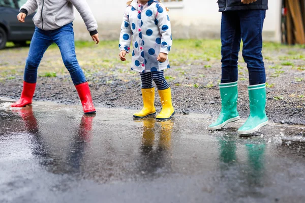 Close-up van drie kinderen, peutermeisje en twee jongens met rode, gele en groene regenlaarzen en wandelen tijdens de sleet. Gelukkige broers en zussen die in een plas springen. Plezier hebben buiten, actief gezin — Stockfoto