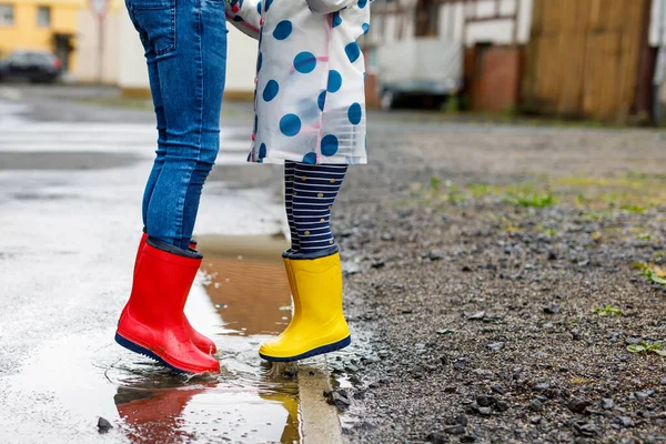 Detailní záběr dvou dětí, batolat a chlapečka v červených a žlutých holínkách na dešti, jak chodí během plískanice. Šťastní sourozenci, bratr a sestra skáčou do kaluže. Bavíte se venku, aktivní rodina — Stock fotografie