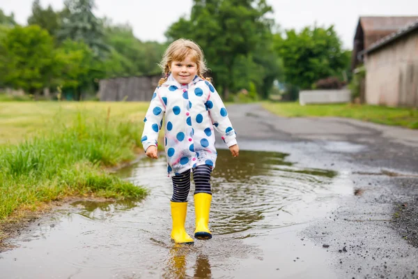 Niña pequeña con botas de lluvia amarillas, corriendo y caminando durante el aguanieve en el día nublado lluvioso. Lindo niño feliz en ropa colorida saltando en el charco, salpicaduras de agua, actividad al aire libre —  Fotos de Stock