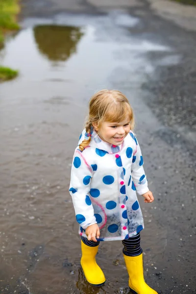 Petite fille en bas âge portant des bottes de pluie jaunes, courant et marchant pendant la neige fondante par temps de pluie nuageux. Enfant heureux mignon en vêtements colorés sautant dans la flaque d'eau, éclaboussures d'eau, activité de plein air — Photo