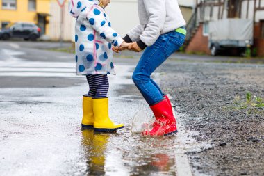 Kırmızı ve sarı çizme giyen, yağmurluk içinde yürüyen iki çocuk, yeni yürümeye başlayan kız ve çocuk. Mutlu kardeşler, kız ve erkek kardeşler gölete atlıyor. Dışarıda eğleniyor, aktif bir aile.