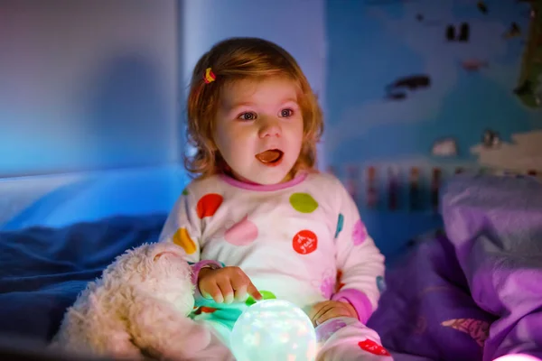 Menina bonito pequena criança brincando com lâmpada de luz colorida noite antes de ir para a cama. Dorminhoco cansado bebê filha em roupa de noite se divertindo. Criança saudável não dorme . — Fotografia de Stock