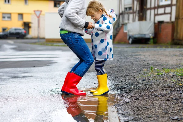 Detailní záběr dvou dětí, batolat a chlapečka v červených a žlutých holínkách na dešti, jak chodí během plískanice. Šťastní sourozenci, bratr a sestra skáčou do kaluže. Bavíte se venku, aktivní rodina — Stock fotografie