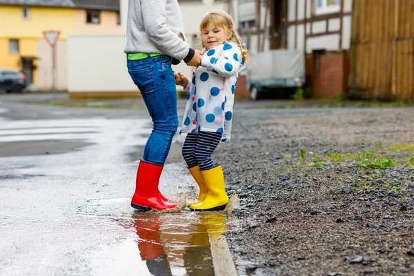 Detailní záběr dvou dětí, batolat a chlapečka v červených a žlutých holínkách na dešti, jak chodí během plískanice. Šťastní sourozenci, bratr a sestra skáčou do kaluže. Bavíte se venku, aktivní rodina — Stock fotografie