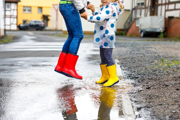 Großaufnahme von zwei Kindern, einem kleinen Mädchen und einem kleinen Jungen, die rote und gelbe Regenstiefel tragen und bei Schneeregen spazieren gehen. Glückliche Geschwister, Bruder und Schwester springen in die Pfütze. Spaß im Freien, aktive Familie — Stockfoto