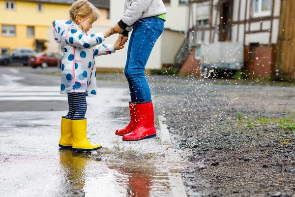 两个小孩的特写镜头，一个是蹒跚学步的小女孩，另一个是穿着红色和黄色雨靴的小男孩，他们在雨夹雪中散步。快乐的兄弟姐妹，兄妹跳进了水坑。户外运动，活跃的家庭 — 图库照片