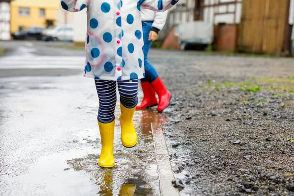 Großaufnahme von zwei Kindern, einem kleinen Mädchen und einem kleinen Jungen, die rote und gelbe Regenstiefel tragen und bei Schneeregen spazieren gehen. Glückliche Geschwister, Bruder und Schwester springen in die Pfütze. Spaß im Freien, aktive Familie — Stockfoto