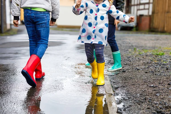 三个小孩，一个是蹒跚学步的小女孩，另一个是穿着红、黄、绿雨靴，在雨夹雪中走路的小孩。快乐的兄弟姐妹跳进了水坑。户外运动，活跃的家庭 — 图库照片