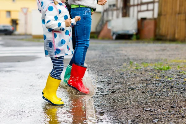 Großaufnahme von drei Kindern, einem kleinen Mädchen und zwei kleinen Jungen, die rote, gelbe und grüne Regenstiefel tragen und bei Schneeregen laufen. Glückliche Geschwister springen in eine Pfütze. Spaß im Freien, aktive Familie — Stockfoto