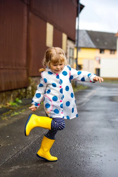 Menina pequena usando botas de chuva amarelas, correndo e andando durante o trenó em dia nublado chuvoso. Criança feliz bonito em roupas coloridas pulando em poça, salpicando com água, atividade ao ar livre — Fotografia de Stock