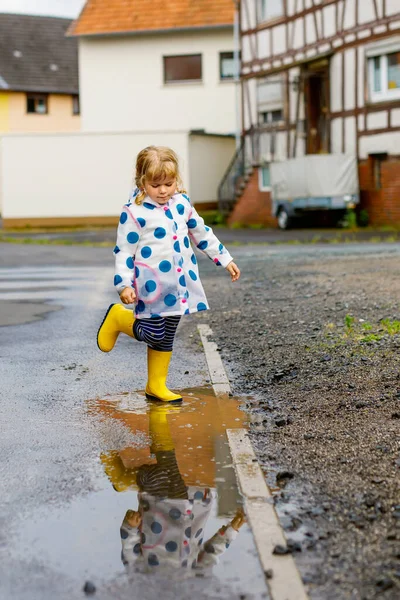 Küçük kız sarı çizme giyiyor, yağmurlu bir günde yağmurda yağmurda koşuyor ve yürüyor. Renkli giysiler içinde şirin mutlu bir çocuk su birikintisine atlıyor, su sıçrıyor, açık hava aktivitesi yapıyor. — Stok fotoğraf