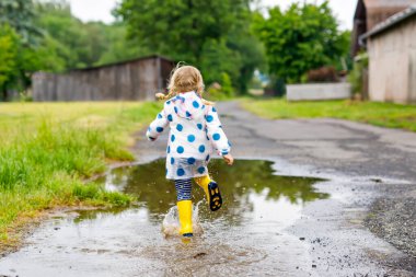 Küçük kız sarı çizme giyiyor, yağmurlu bir günde yağmurda yağmurda koşuyor ve yürüyor. Renkli giysiler içinde şirin mutlu bir çocuk su birikintisine atlıyor, su sıçrıyor, açık hava aktivitesi yapıyor.